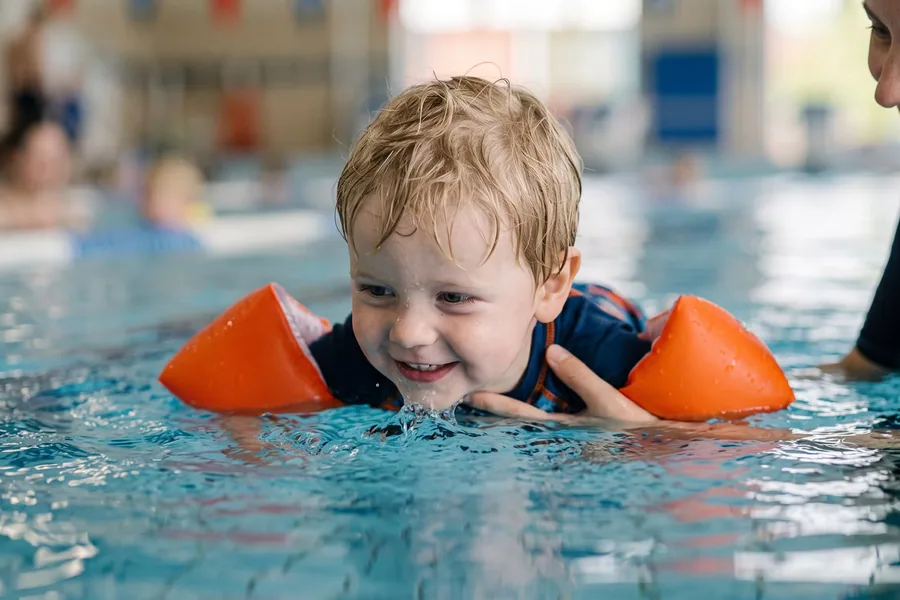 Un bambino con i braccioli avanza a pelo d’acqua, accompagnato dall’istruttrice, durante una lezione genitore-bambino in vasca coperta — Genitore-bambino (2-4 anni)