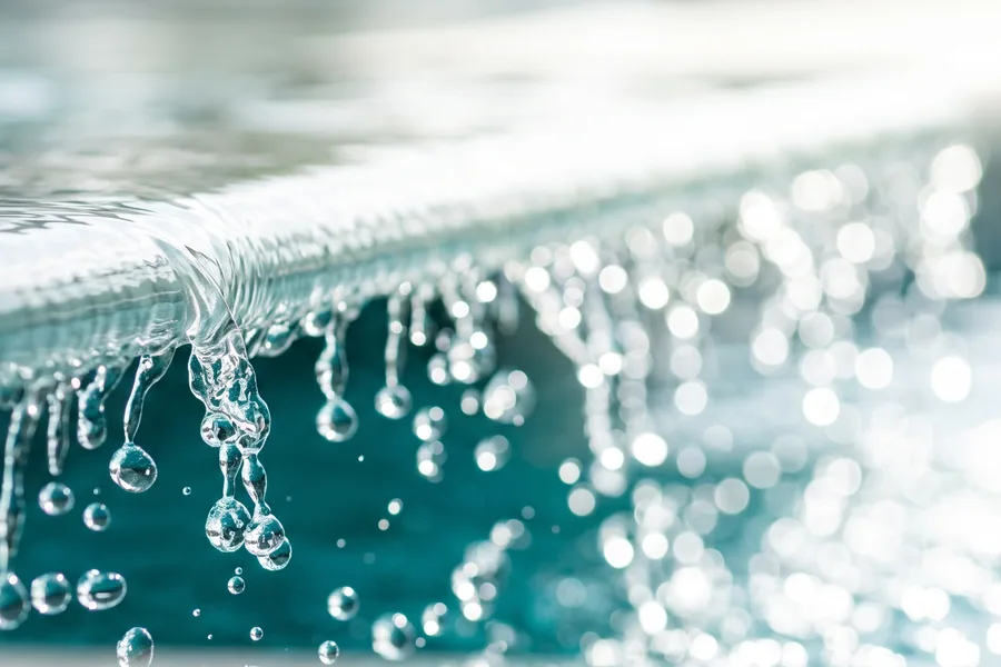 Gocce d’acqua che cadono dal bordo di una vasca, sullo sfondo la superficie illuminata